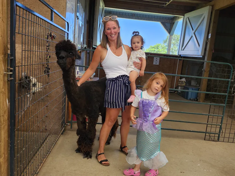 A mother and two children at the barn doorway with a dark alpaca beside them